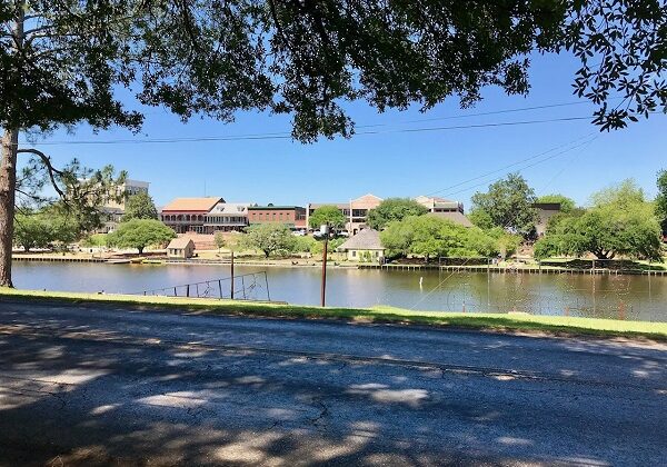 River bank lined with shade trees, a road, and buildings in Natchitoches, Louisiana.